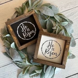 May include: Two wooden signs with the text "He is Risen" in a wreath design. The signs are on a white wood background with a green leafy garland.