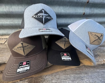May include: Four baseball caps with a mesh back and a diamond-shaped patch with a sword and cross design. The patches are brown, black, and gray. The caps are gray, brown, black, and blue. The text on the patches reads "REDEEMED".