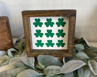 May include: A small, square wooden sign with a dark brown frame. The sign features nine green shamrocks on a white background. The sign is resting on a garland of green leaves.