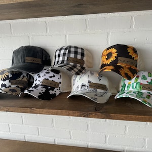 May include: Eight trucker hats with different patterns, including black and white buffalo plaid, sunflowers, and cacti. Each hat has a brown patch sewn on the front that says "mama" in cursive.
