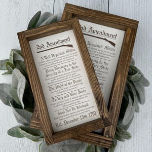 May include: Two framed prints of the 2nd Amendment, featuring the text of the amendment and a rifle illustration. The frames are made of dark brown wood. The prints are displayed on a white surface with green foliage.