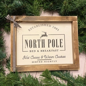 May include: A rustic wooden sign with the text "Established 1901 North Pole Bed & Breakfast Hot Cocoa & Warm Cookies Served Nightly". The sign features a silhouette of a reindeer jumping.