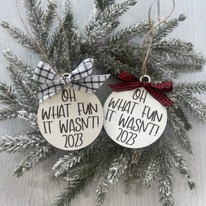 May include: Two white round ornaments with black text that reads "Oh What Fun It Wasn't! 2023". One ornament has a black and white checkered ribbon and the other has a red and white checkered ribbon.