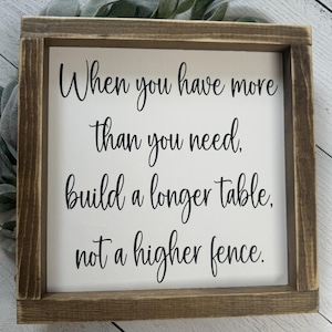 May include: Square wooden sign with a white background and a brown frame. The sign features black cursive text that reads: "When you have more than you need, build a longer table, not a higher fence."