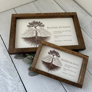 May include: Two framed wooden signs with the text "ROOTED IN FAITH" and an illustration of a tree with a cross. The signs have a dark brown frame and are displayed on a white wooden surface.