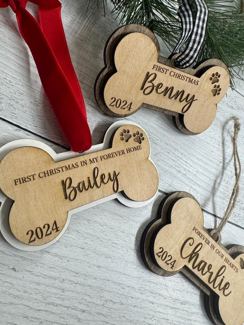 May include: Three wooden dog bone ornaments with the text "First Christmas" and the year "2024" on each. The names "Benny", "Bailey", and "Charlie" are also on the ornaments. The ornament for Charlie has the text "Forever in our hearts".
