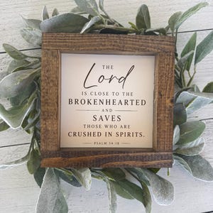 May include: A square wooden sign with the text "The Lord is close to the brokenhearted and saves those who are crushed in spirits. Psalm 34:18." The sign is surrounded by green and grey leaves.