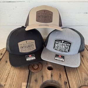 May include: Three trucker hats with a patch that reads "THE RIGHTEOUS MAN WALKS IN HIS INTEGRITY: HIS CHILDREN ARE BLESSED AFTER HIM." The hats are tan, black, and gray with black mesh backs. The hats are on a wooden spool.