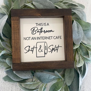 May include: A framed wooden sign with the text "THIS IS A Bathroom NOT AN INTERNET CAFE Shit & Split" in black lettering. The sign is surrounded by a green leafy wreath.