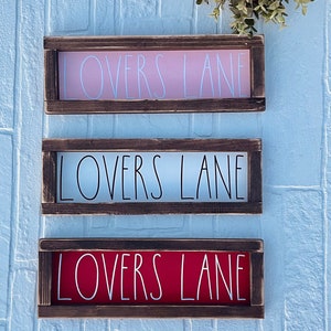 May include: Three wooden signs with the text "LOVERS LANE" in white lettering. The signs are in a brown frame and have different colored backgrounds: pink, white, and red.