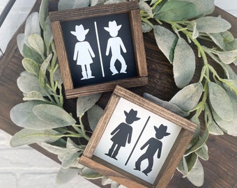 Op de afbeelding: Twee houten borden met een zwart-wit silhouet van een cowboy en een cowgirl. De borden zijn ingelijst in donker hout en hebben een rustieke uitstraling.