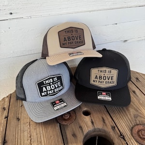 May include: Three trucker hats with the text "THIS IS ABOVE MY PAY GRADE" on a patch. The hats are tan, gray, and black, with mesh backs. The hats are displayed on a wooden spool.
