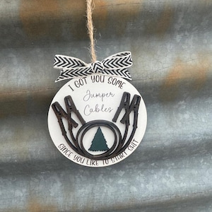 May include: A round, white ornament with the text "I GOT YOU SOME JUMPER CABLES SINCE YOU LIKE TO START SHIT". The ornament features a black and brown design of jumper cables and a green tree. It is topped with a black and white bow.