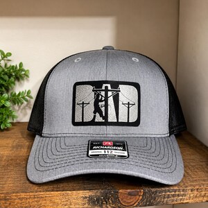 Peut inclure: Casquette de camionneur grise avec un dos en filet noir. L'avant présente un écusson avec le mot "DAD" et une silhouette d'un électricien sur des lignes électriques. La casquette a une visière incurvée et une sangle réglable.