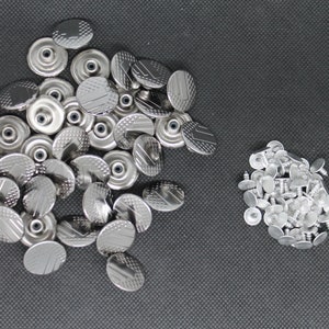 May include: A pile of silver metal snap fasteners, some with a textured surface, and a pile of silver metal rivets.