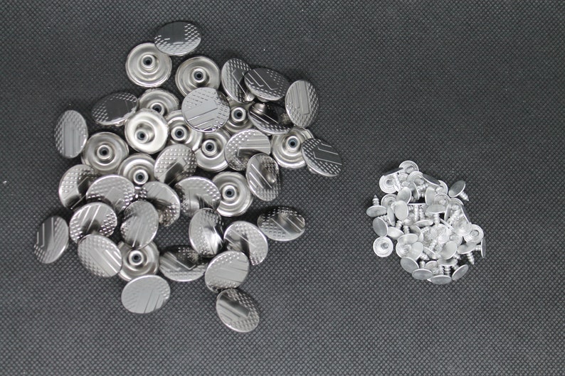 May include: A pile of silver metal snap fasteners, some with a textured surface, and a pile of silver metal rivets.