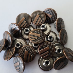 May include: A close-up of a pile of copper-toned metal buttons with a textured surface and a raised design. The buttons have a silver-toned shank on the back.