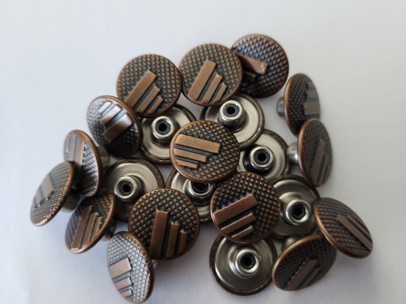 May include: A close-up of a pile of copper-toned metal buttons with a textured surface and a raised design. The buttons have a silver-toned shank on the back.