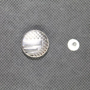 May include: A silver button with a patterned design resembling a flag. The button is next to a smaller, plain silver button.