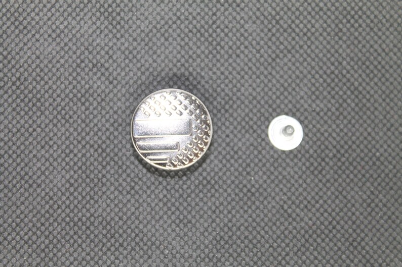 May include: A silver button with a patterned design resembling a flag. The button is next to a smaller, plain silver button.