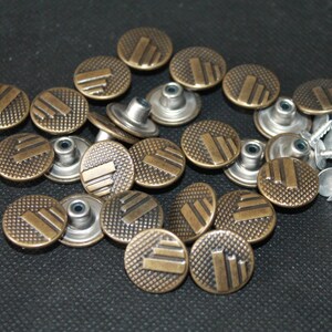 May include: A close-up of a group of 18 antique gold metal buttons with a decorative pattern. The buttons have a raised design and are arranged in a circular pattern. There are also 15 silver metal rivets in the image.