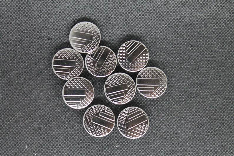 May include: A group of nine silver-colored watch batteries with a patterned design on the top.