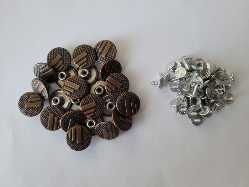 May include: A close-up of two piles of metal buttons. One pile is made up of 18 copper-colored buttons with a raised design. The other pile is made up of 25 silver-colored buttons with a flat top.