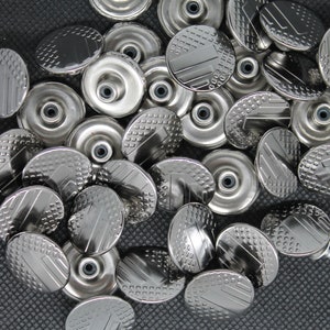 May include: A close-up of a pile of silver metal buttons with a textured design. The buttons are round and have a raised center.