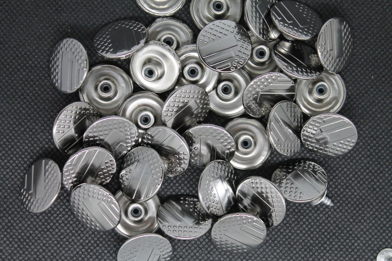 May include: A close-up of a pile of silver metal buttons with a textured design. The buttons are round and have a raised center.