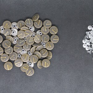 May include: A large pile of gold-colored metal buttons with a textured design. There is a smaller pile of silver-colored metal buttons to the right.