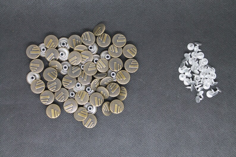 May include: A large pile of gold-colored metal buttons with a textured design. There is a smaller pile of silver-colored metal buttons to the right.