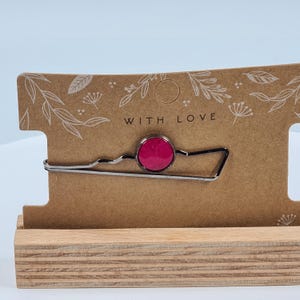 Könnte beinhalten: Eine silberfarbene Krawattenklammer mit einem runden, magenta-farbenen Stein. Die Krawattenklammer wird auf einer braunen Karte mit dem Schriftzug "WITH LOVE" und floralen Designs präsentiert. Die Karte steht auf einem Holzständer.