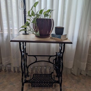 Peut inclure: Une table d'appoint avec un plateau rectangulaire en bois et une base en métal noir issue d'un cadre de machine à coudre vintage. La table supporte un pot en céramique violet foncé avec une plante pothos, une bougie noire et deux livres.