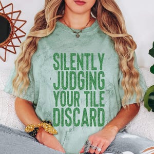 May include: A light green acid-wash t-shirt featuring the dark green text "SILENTLY JUDGING YOUR TILE DISCARD" printed in a bold, sans-serif font across the chest. The shirt has short sleeves and a crew neckline, showcasing a textured, mottled finish.