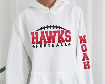 May include: White hoodie with a red glitter design that says "HAWKS FOOTBALL" with a football graphic above the text. The right sleeve has the name "NOAH" in red glitter.
