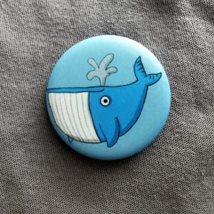 May include: A circular pinback button featuring a cartoon illustration of a blue whale on a light blue background. The whale has a white underbelly with vertical lines, a single visible eye, and a grey water spout emerging from its blowhole.