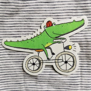 May include: An embroidered patch featuring a bright green crocodile wearing a small red hat whilst riding a bicycle. The bicycle has a yellow headlight and thin black wheels. The patch is set against a background of white fabric with thin blue stripes.