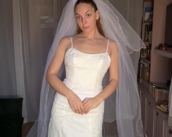 Puede incluir: Un vestido de novia blanco hasta el suelo con tirantes finos y un corpiño ajustado con intrincados bordados de encaje. El vestido se combina con un velo largo de tul transparente y superpuesto que cae sobre los hombros.