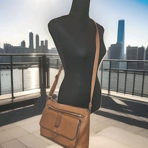 May include: A brown leather crossbody bag with a long adjustable strap. The bag has a flap pocket on the front and a zippered pocket on the top. The bag is displayed on a black mannequin against a city skyline background.
