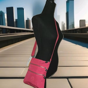 May include: A bright pink crossbody bag with three zippered compartments and a long, thin strap. The bag is displayed on a black mannequin against a backdrop of a city skyline. The bag has a small, circular logo on the front.