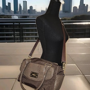 May include: A brown handbag with a snakeskin pattern on the front flap and side pockets. The bag has a long, adjustable strap and a shorter handle. The bag is displayed on a black mannequin.