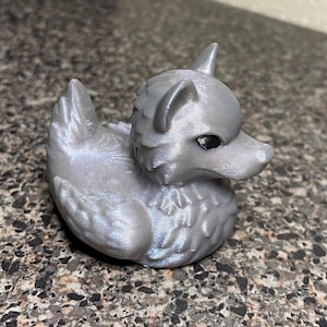 May include: A silver-colored rubber duck with a dragon-like head and black eyes. The duck is sitting on a textured surface.