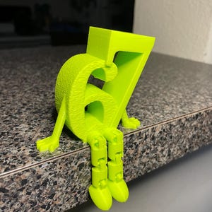 May include: A bright neon green number seven figure with articulated legs and arms, sitting on a countertop. The number has a textured surface and is 3D-printed. The figure is a whimsical decorative item.