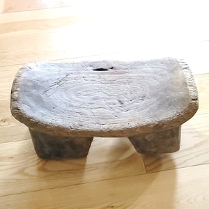 May include: A rustic wooden stool with a rounded top and four short legs. The stool is made of dark wood with a natural finish and shows signs of wear and tear.