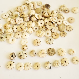 May include: A collection of white, carved, shell buttons with a flower design. The buttons are arranged in a pile on a white surface.
