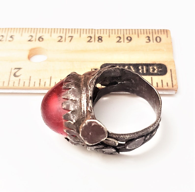 Puede incluir: Un anillo de plata con una piedra de vidrio roja engastada en el centro. El anillo est&aacute; decorado con peque&ntilde;as piezas de plata y tiene una banda texturizada.