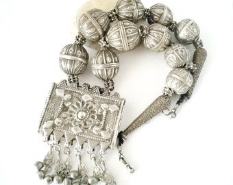 May include: A silver necklace featuring a large, ornate pendant and multiple round beads. The pendant is square-shaped with intricate designs and a floral motif. The necklace has a long, thin chain with a clasp.