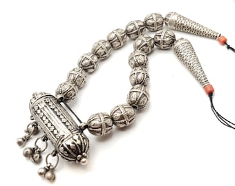 May include: A silver necklace featuring a cylindrical pendant and a series of round beads. The pendant is adorned with intricate designs and is attached to the necklace by a small chain. The beads are also decorated with intricate patterns.