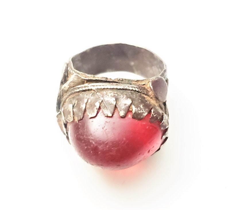 Puede incluir: Un anillo de plata con una piedra preciosa roja engastada en un engaste de plata decorativo. El anillo tiene una banda texturizada y detalles intrincados.