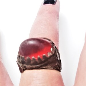 Puede incluir: Un anillo de plata con una piedra preciosa roja engastada en un engaste decorativo y dentado. El anillo se lleva en un dedo.